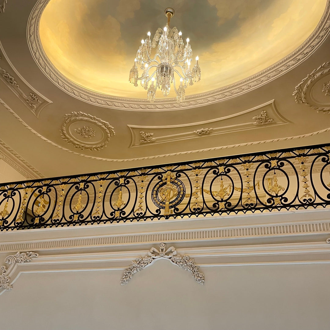 Ornate black and gold wrought iron handrail by Choufani, set against a classical interior with intricate ceiling moldings, a painted dome, and a crystal chandelier.