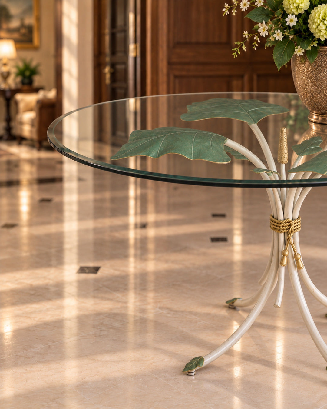 A round glass-top table with a sculptural white metal base resembling tropical leaves, tied together with rope, shown in a bright, sunlit interior hallway.