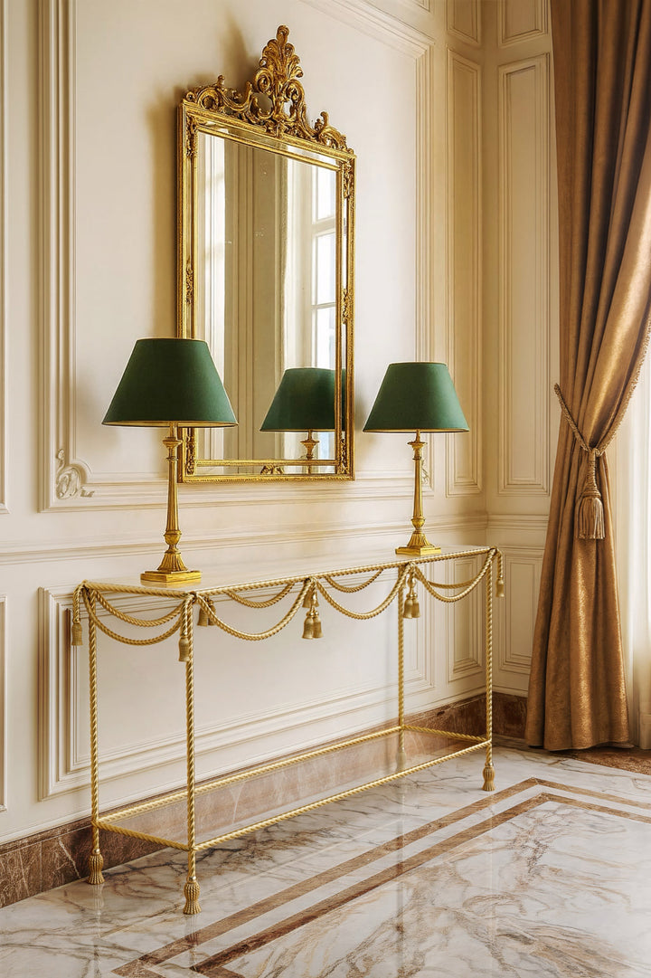 Allegra Console Table in hand-finished gold, styled in a classic interior with rope-tassel detailing, a gilded mirror, and twin green-shaded lamps on a polished marble floor.