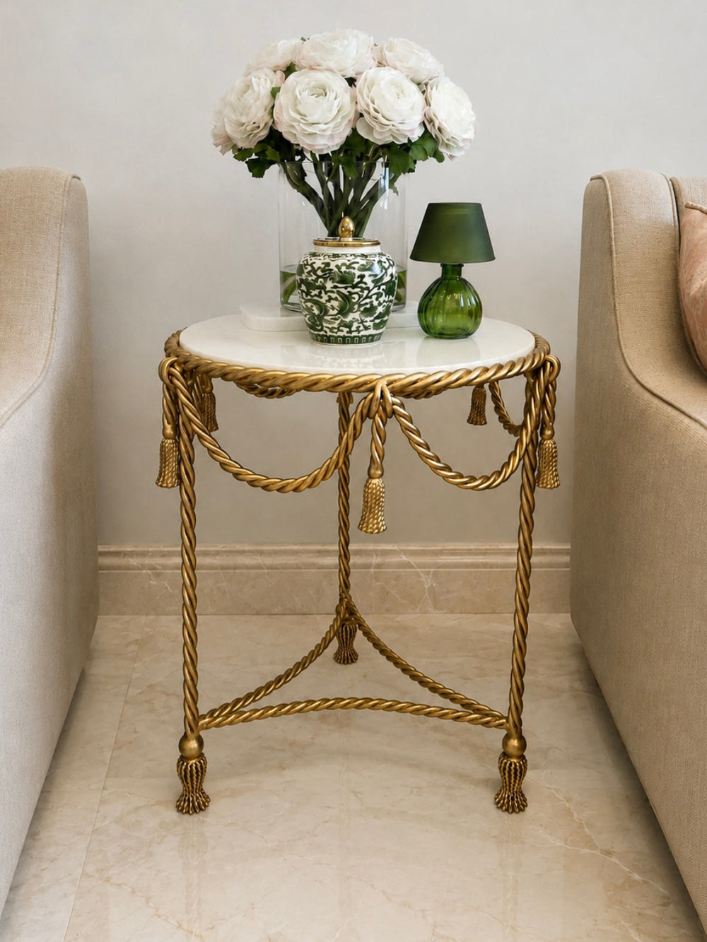 A decorative round side table with a white marble top and ornate gold rope-style metal frame, styled between two beige armchairs. The table holds a vase of white roses and a small green table lamp.