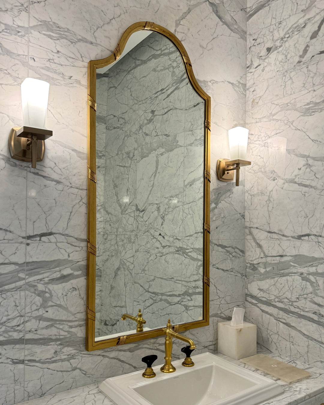Full view of the Audrey Mirror with an arched crown and antique gold frame, mounted above a marble sink with brass fixtures.