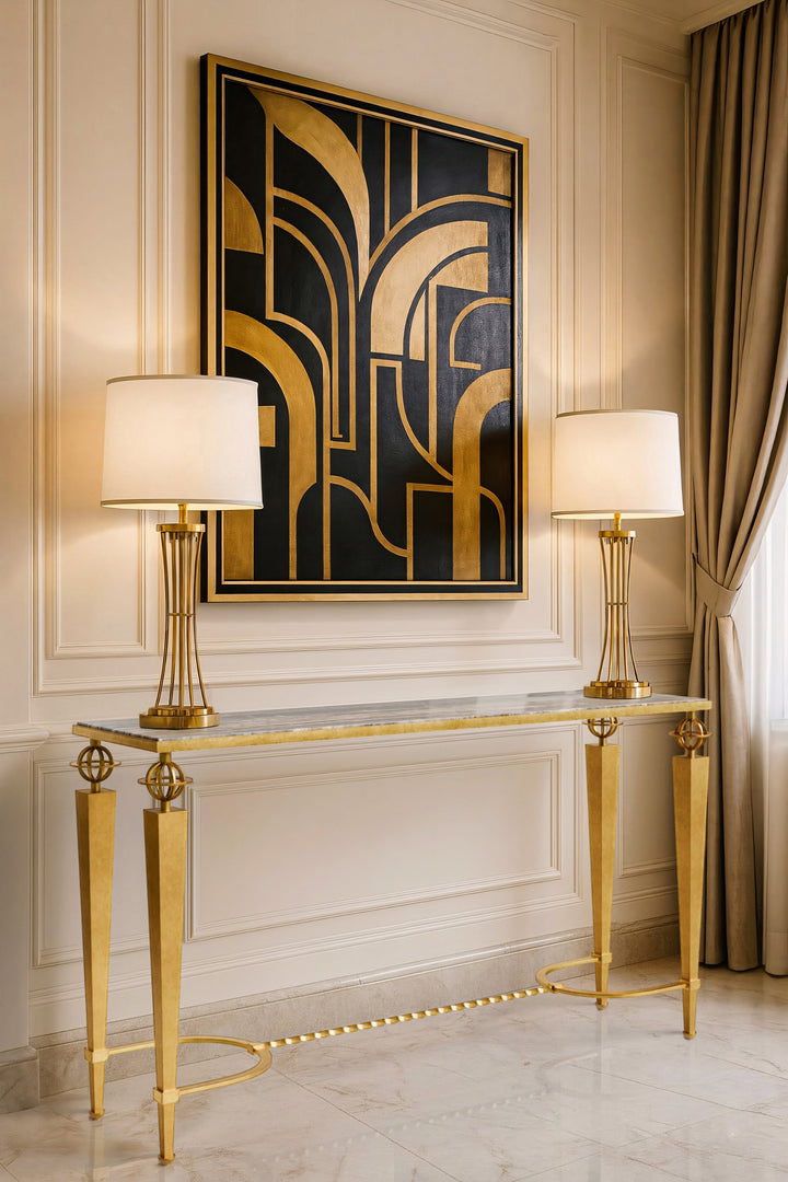 Gilbert console table with tapered gold legs, marble top, and Art Deco detailing, styled with gold table lamps and geometric black-and-gold artwork in an elegant cream interior.