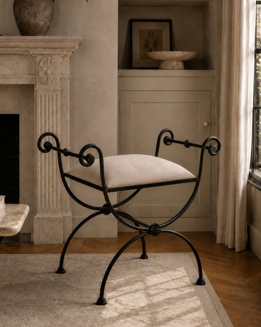 A black wrought-iron stool with a curved scrollwork frame and a off-white upholstered seat cushion, placed in a softly lit room with a fireplace, built-in cabinet, rug, and curtains.
