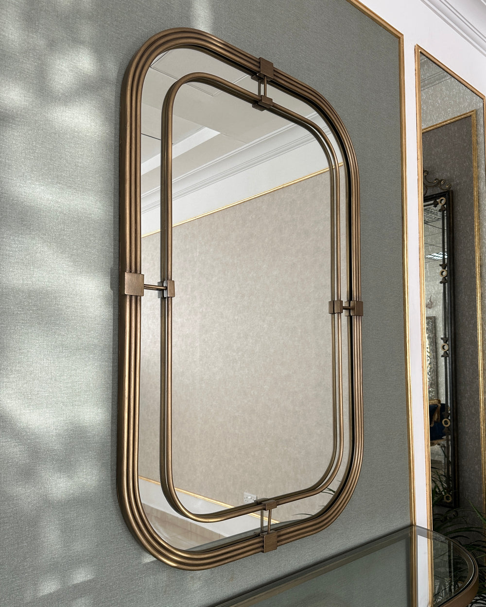 Elegant rectangular wall mirror with rounded edges and a double bronze frame, mounted on a textured wall panel with gold trim beside a glass-topped console.